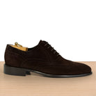 A dark brown suede wingtip oxford shoe with brogue detailing, displayed on a wooden surface against a white background.