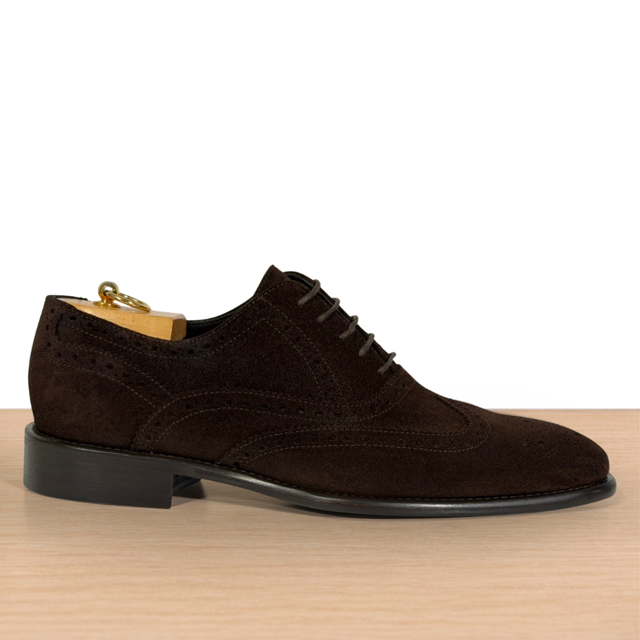 A dark brown suede wingtip oxford shoe with brogue detailing, displayed on a wooden surface against a white background.