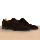 A pair of dark brown suede wingtip oxford shoes with brogue detailing, displayed on a light wood surface against a white background.