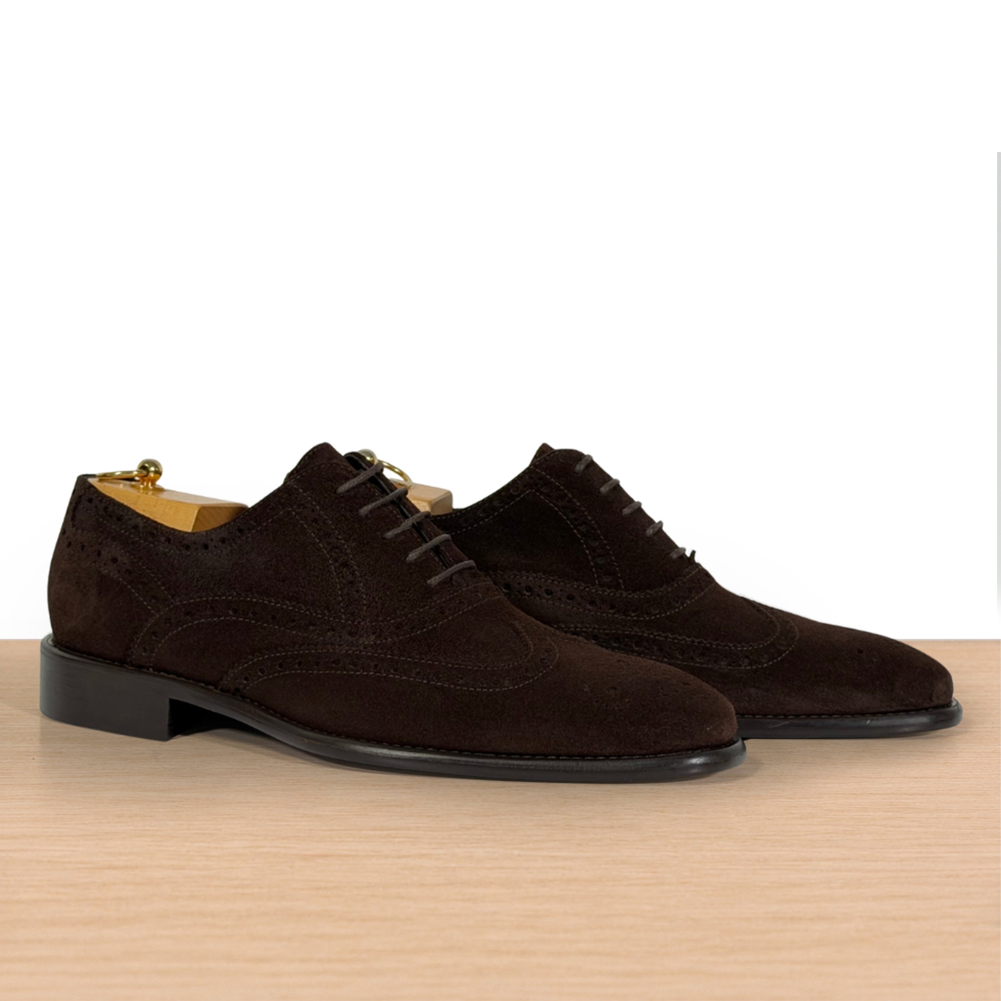 A pair of dark brown suede wingtip oxford shoes with brogue detailing, displayed on a light wood surface against a white background.