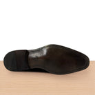 The outsole of a dark brown leather dress shoe, showing the V-shaped center strip and heel cap, resting on a light wood surface against a white background.