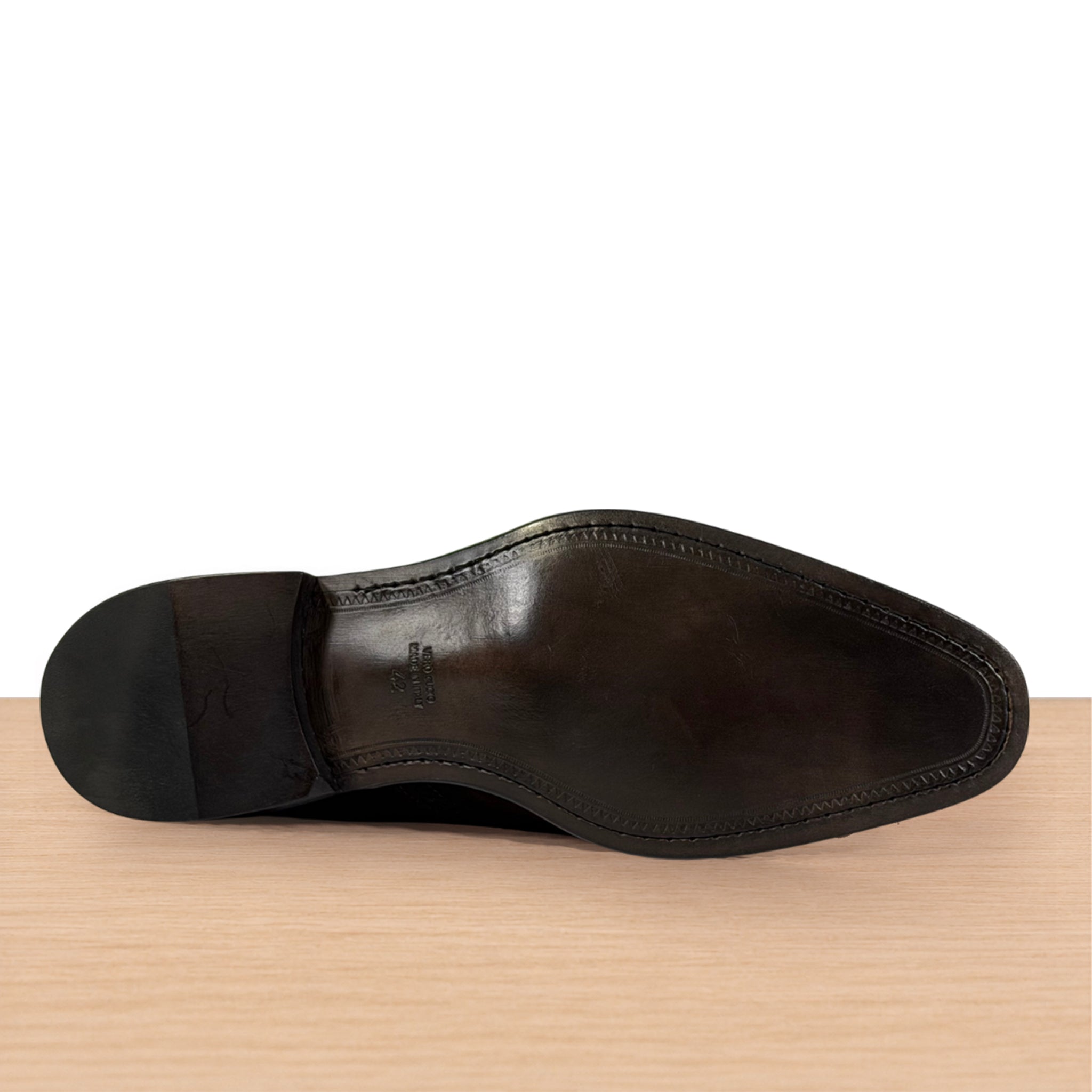 The outsole of a dark brown leather dress shoe, showing the V-shaped center strip and heel cap, resting on a light wood surface against a white background.