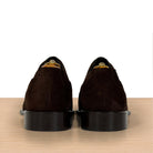 A pair of dark brown suede oxford shoes shown from the back, resting on a light wood surface against a white background.