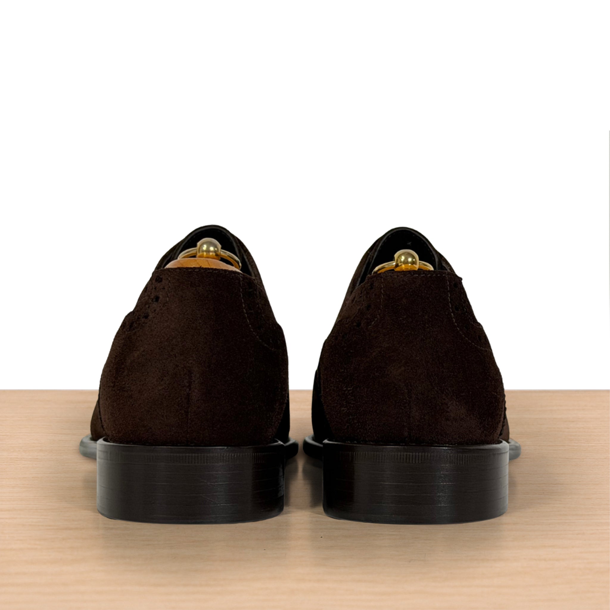 A pair of dark brown suede oxford shoes shown from the back, resting on a light wood surface against a white background.