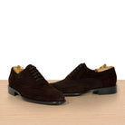 A pair of dark brown suede wingtip oxford shoes with black laces, displayed on a light wood surface against a white background.