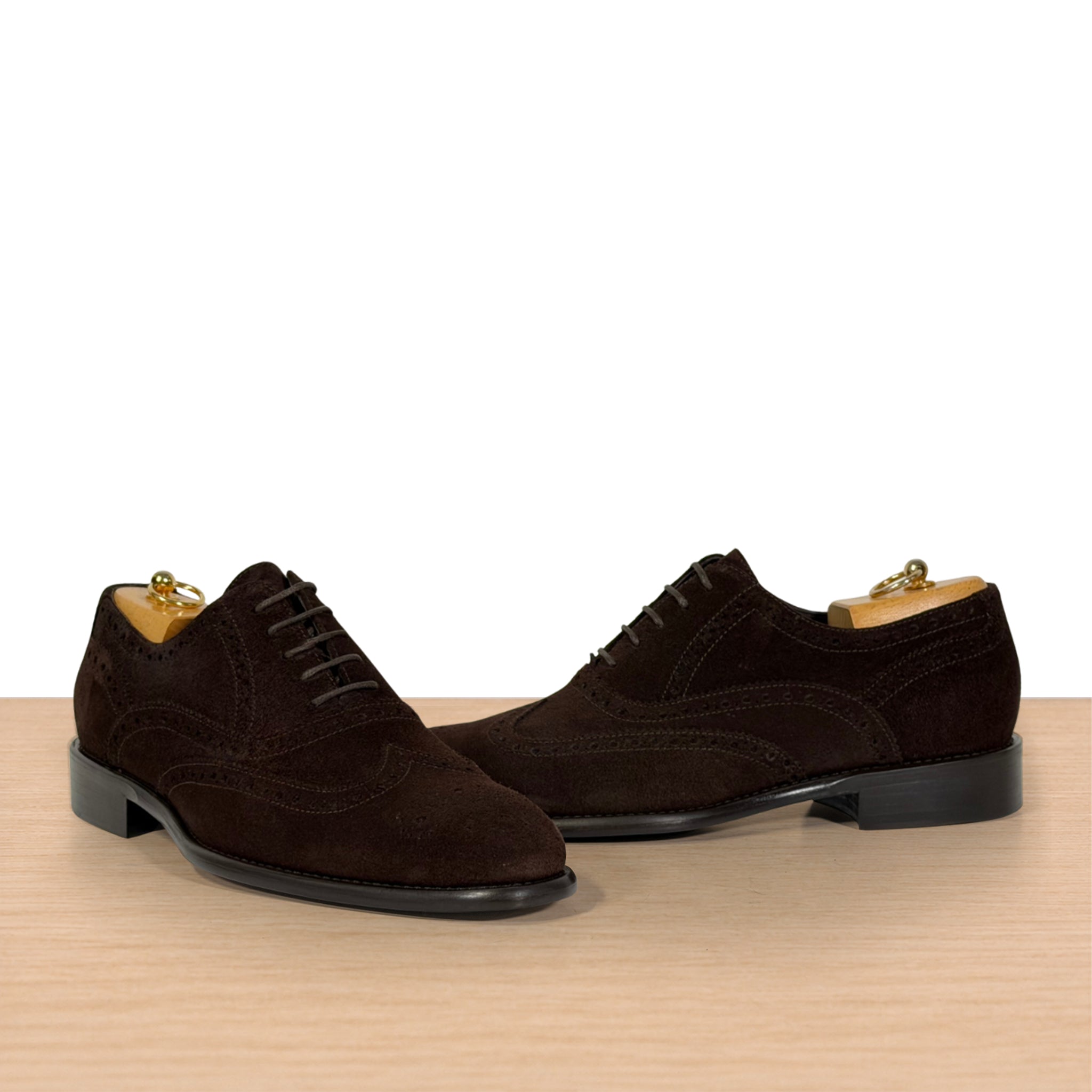A pair of dark brown suede wingtip oxford shoes with black laces, displayed on a light wood surface against a white background.