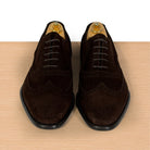 A pair of dark brown suede wingtip oxford shoes with laces and black soles, displayed on a light wood surface against a white background.