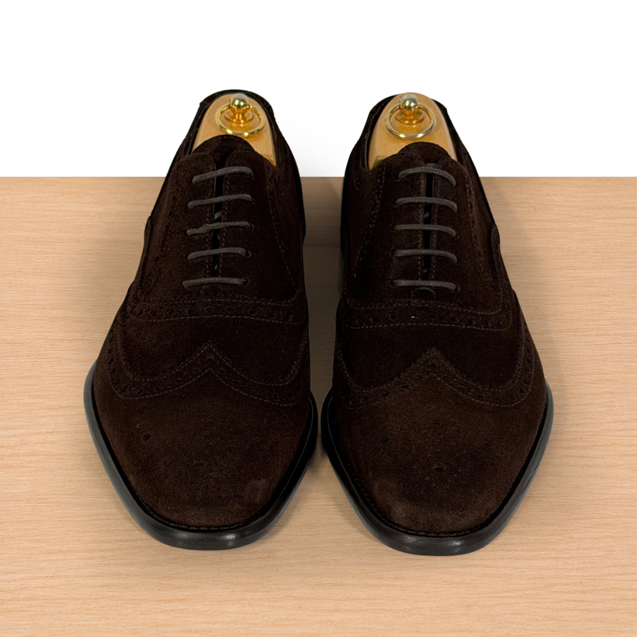 A pair of dark brown suede wingtip oxford shoes with laces and black soles, displayed on a light wood surface against a white background.