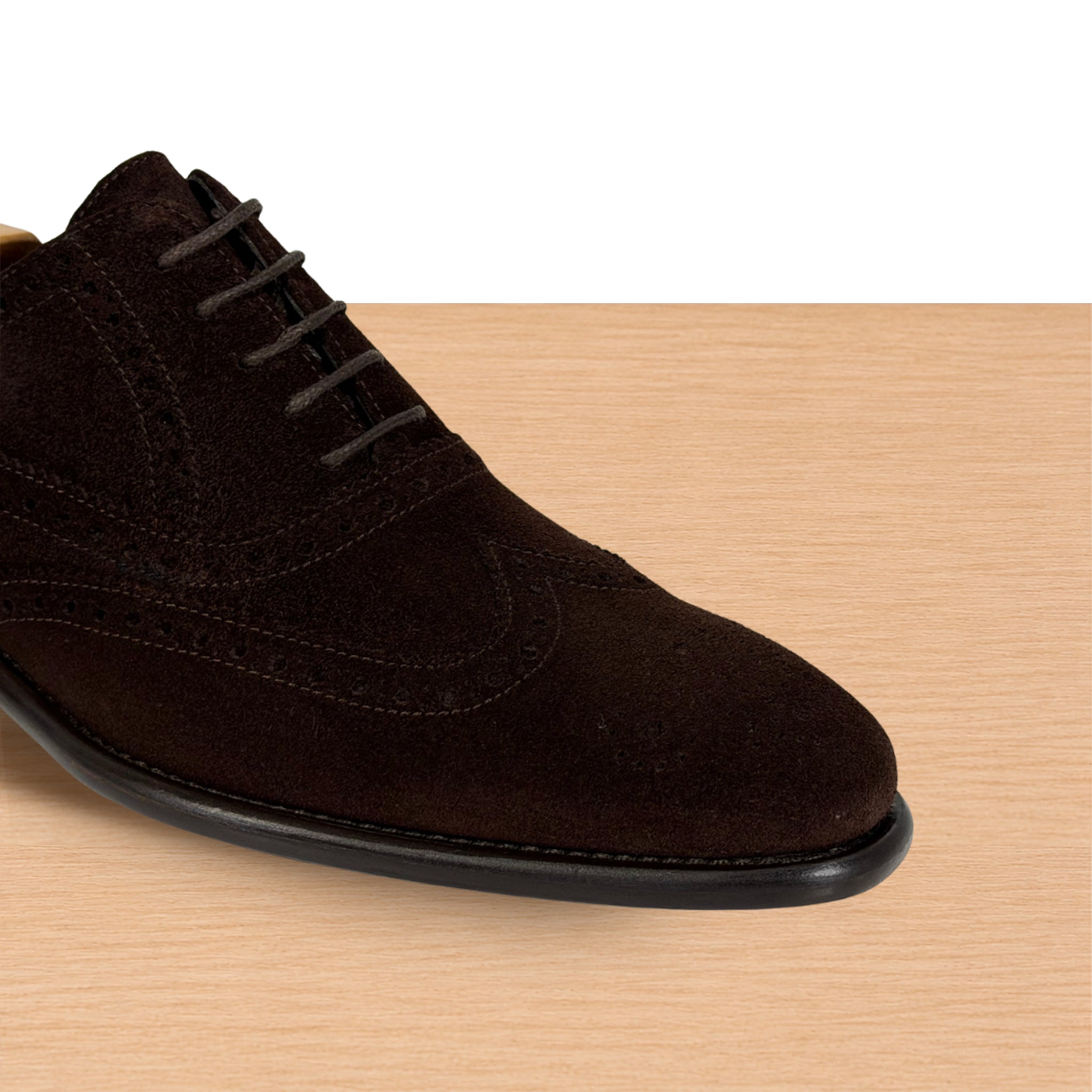 A dark brown suede lace-up dress shoe shown from a side angle on a light wood surface with a white background above.