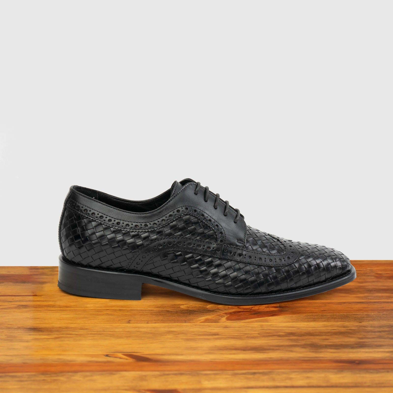 Black woven dress shoe on a wooden surface with a light gray background