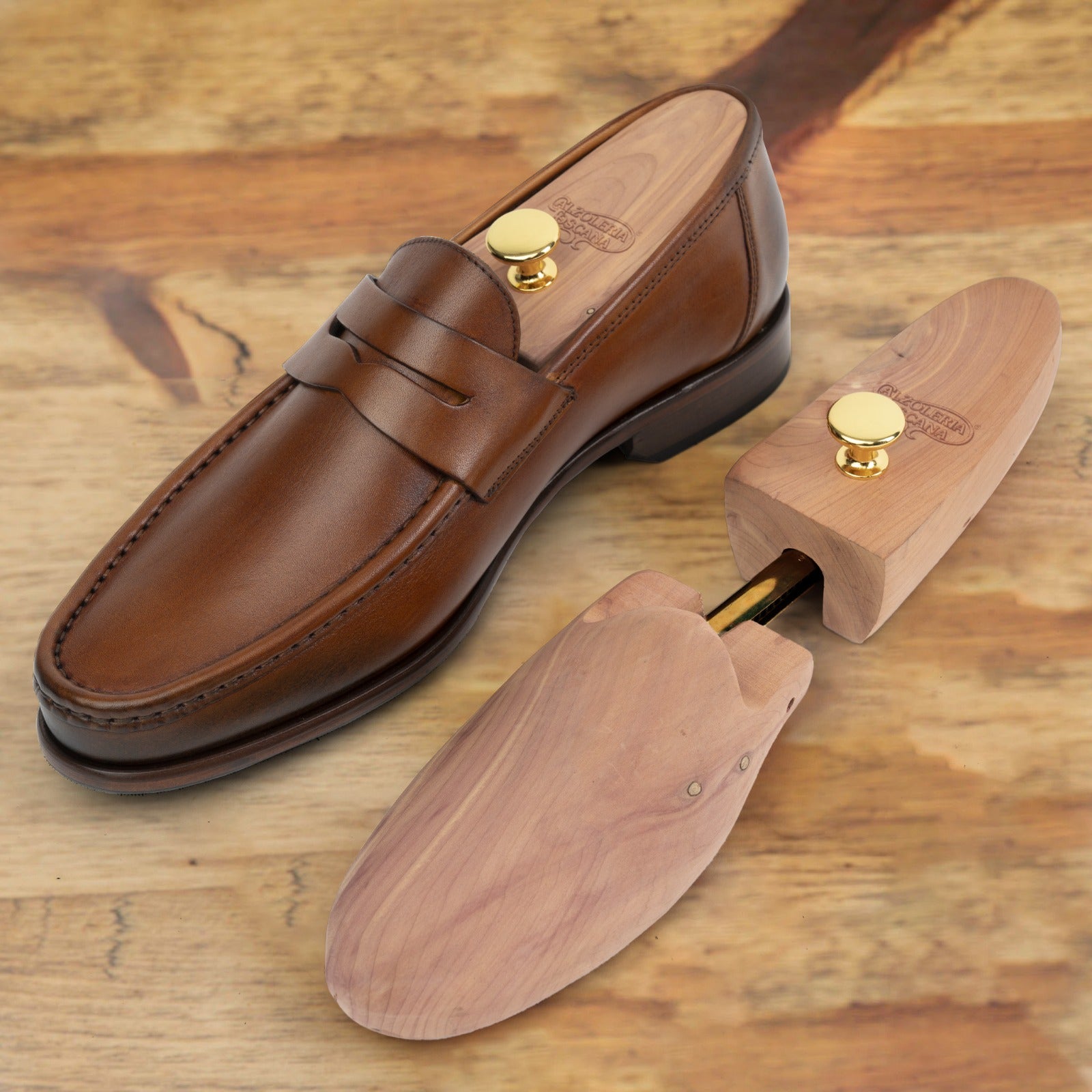 Brown leather penny loafer with a wooden shoe tree inserted, alongside a second shoe tree, displayed on a wooden surface.