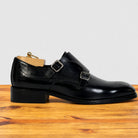A black leather double strap monk shoe with silver-tone buckles, displayed in side profile on a wooden surface against a light gray background.