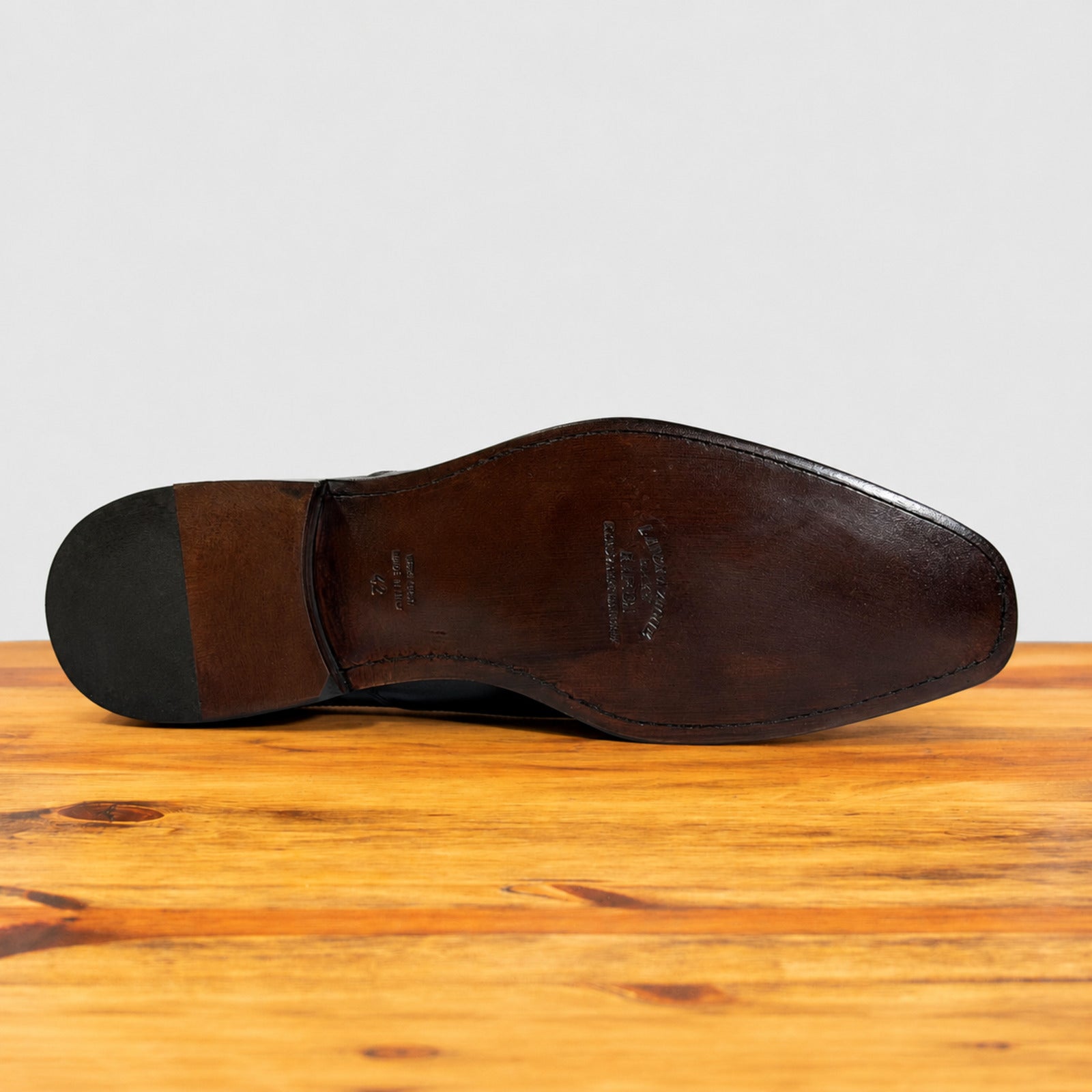 The outsole of a leather dress shoe is shown from a top-down view, resting on a wooden surface against a light gray background.