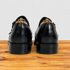 A pair of black leather lace-up dress shoes shown from the back, resting on a wooden surface against a light gray background.