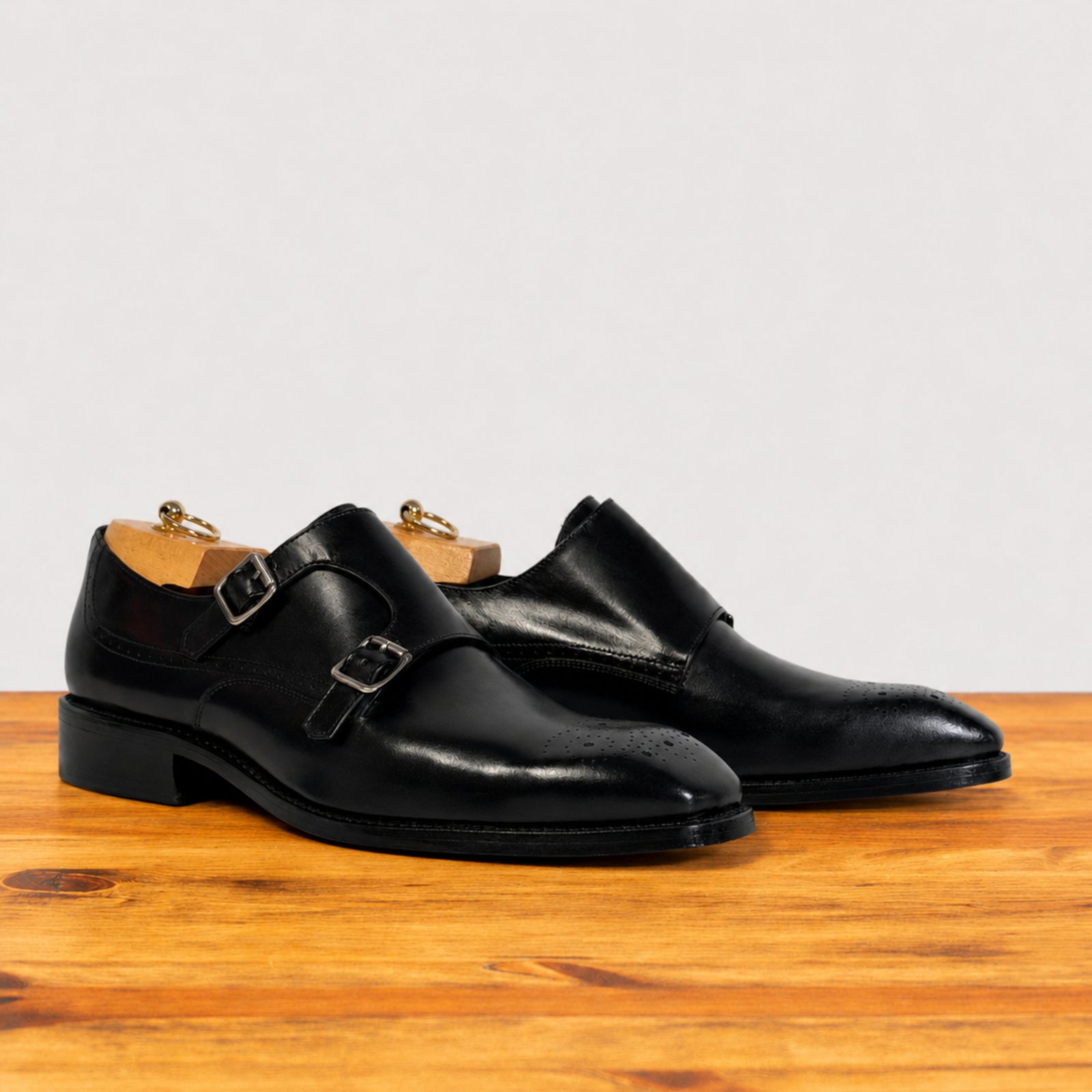 A pair of black leather double strap monk-style dress shoes with brogue detailing, displayed on wooden shoe trees on a wooden surface against a light gray background.