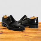 A pair of black double-breasted monk strap dress shoes with brogue detailing, over a polished wooden surface against a light gray background.