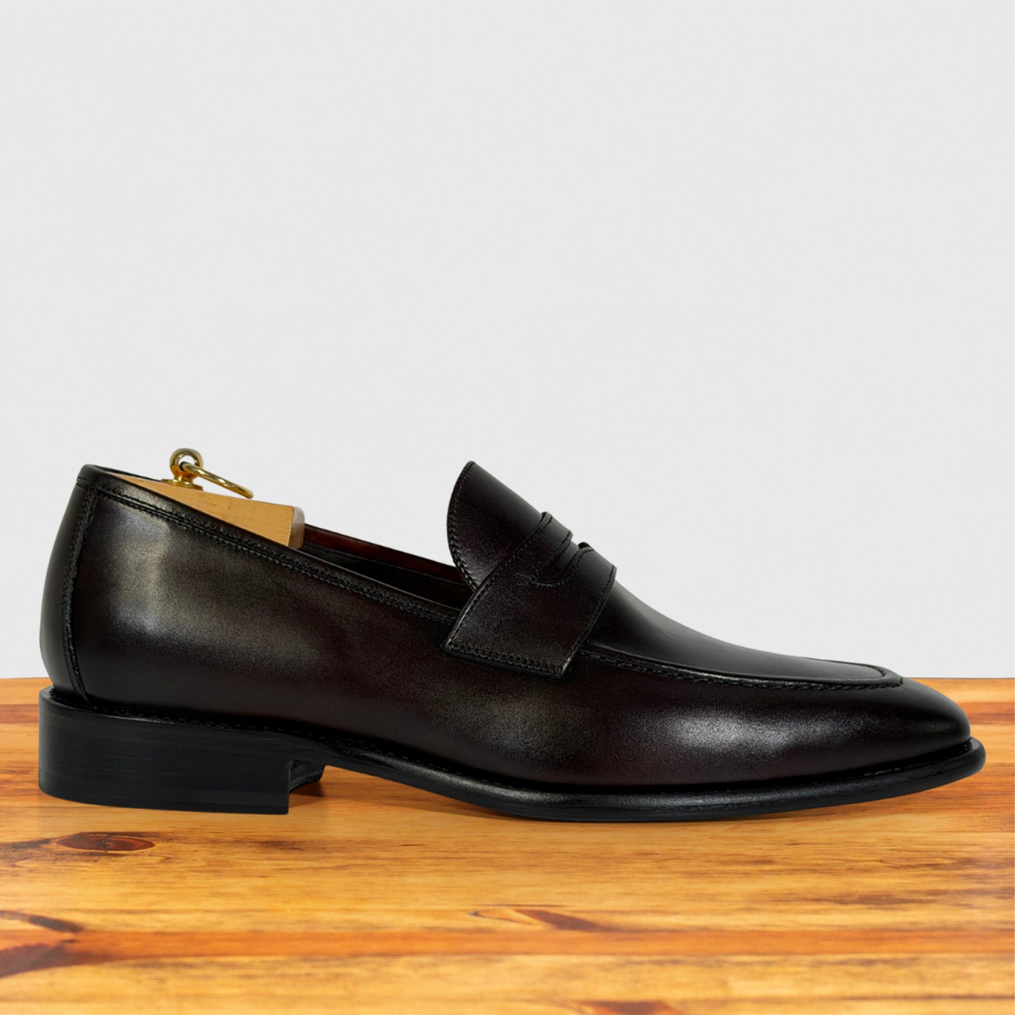 A Burgundy leather penny loafer shoe with a low stacked heel, displayed on a wooden surface against a light gray background.