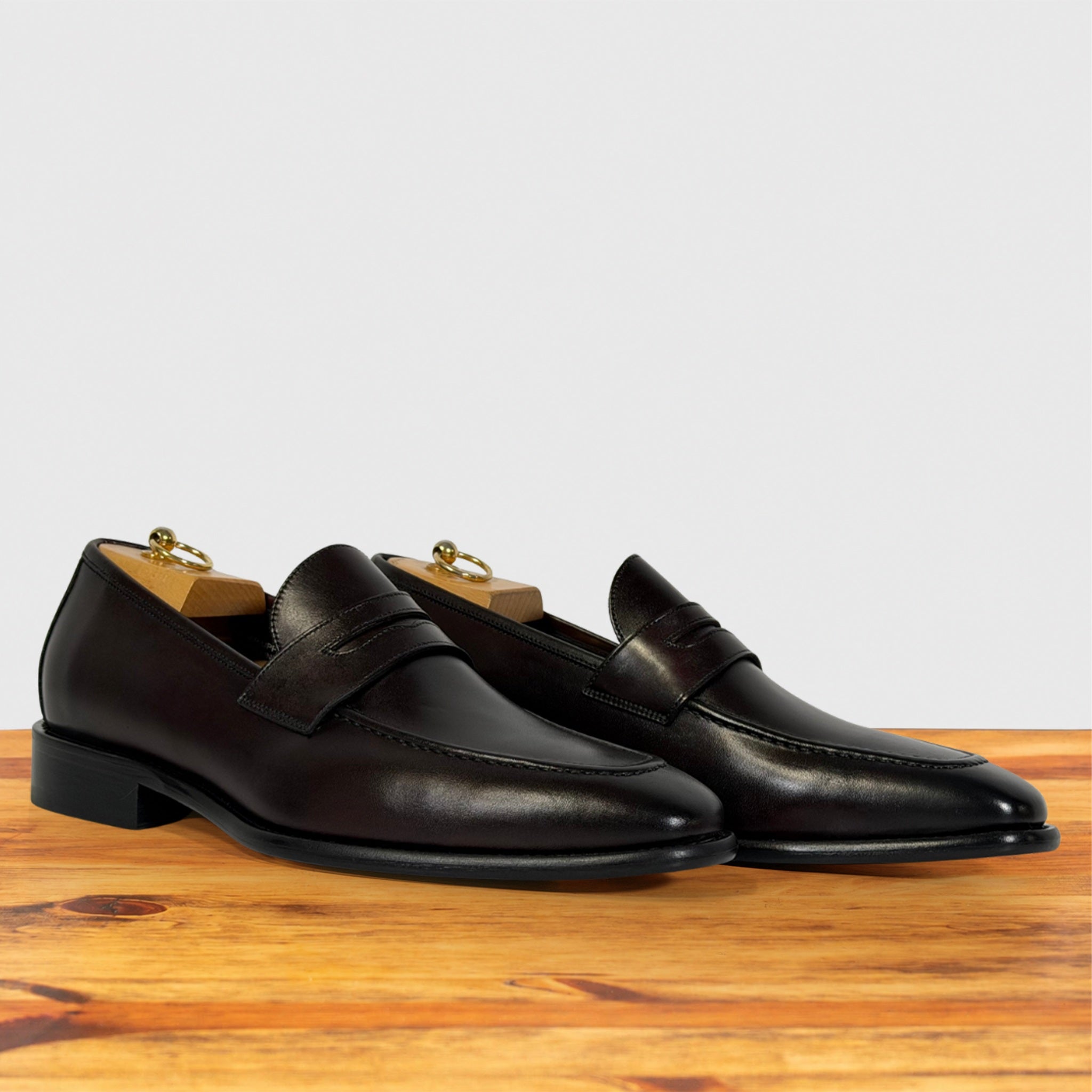 A pair of Burgundy leather penny loafers with low block heels, displayed on wooden shoe trees on a wooden surface against a light gray background.