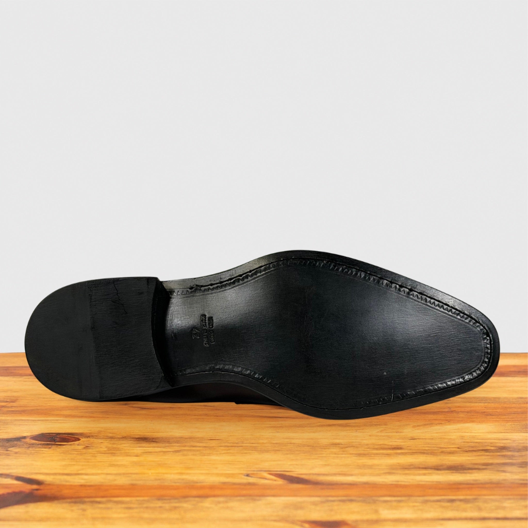 The black leather sole of a dress shoe, shown from the bottom view, resting on a wooden surface against a light gray background.