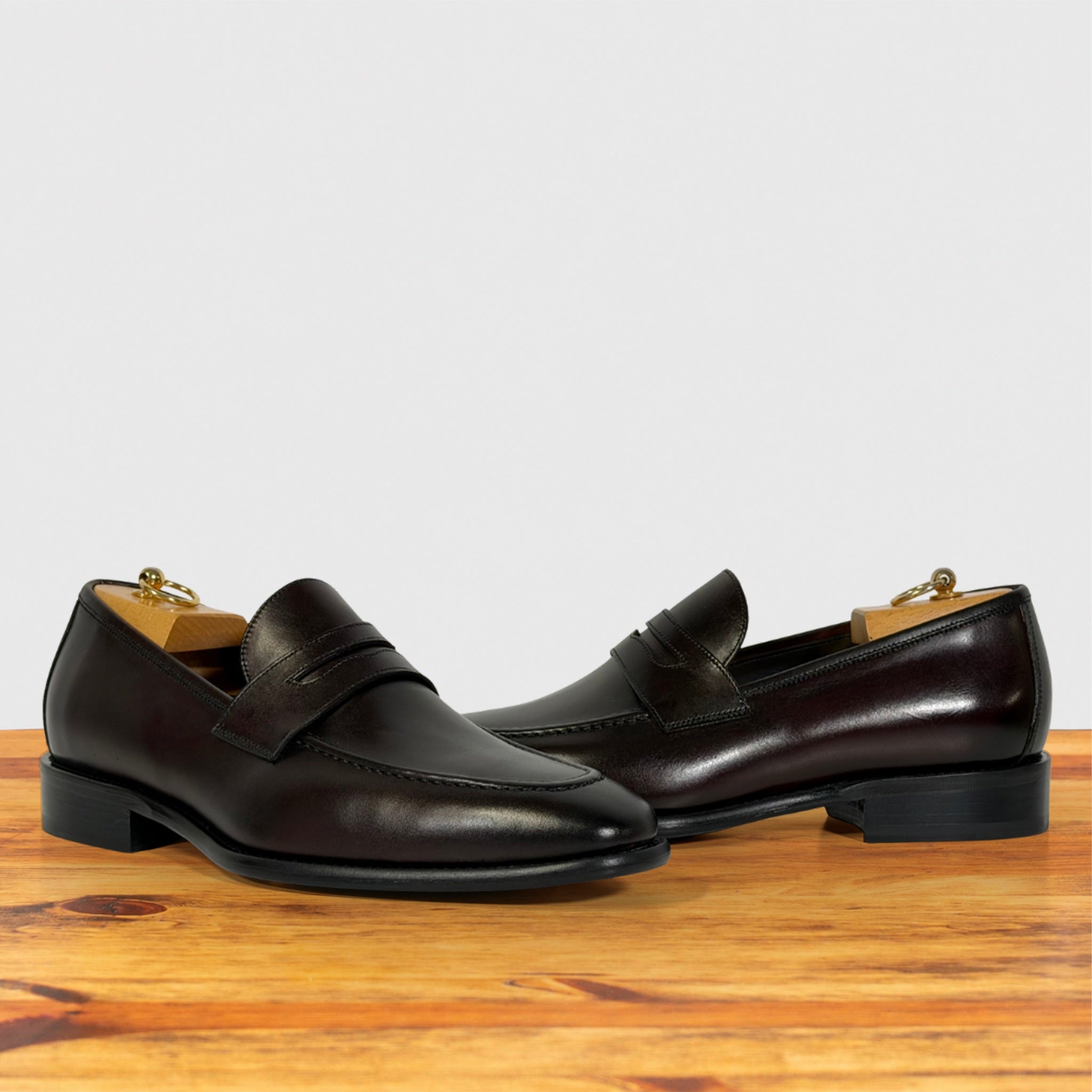 A pair of Burgundy leather penny loafers with black soles, on a wooden surface against a light gray background.