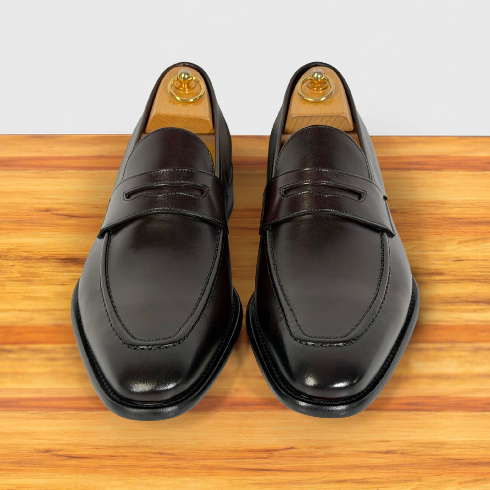 A pair of men's Burgundy leather penny loafers with a glossy finish, displayed on a wooden surface with wooden shoe trees visible inside.