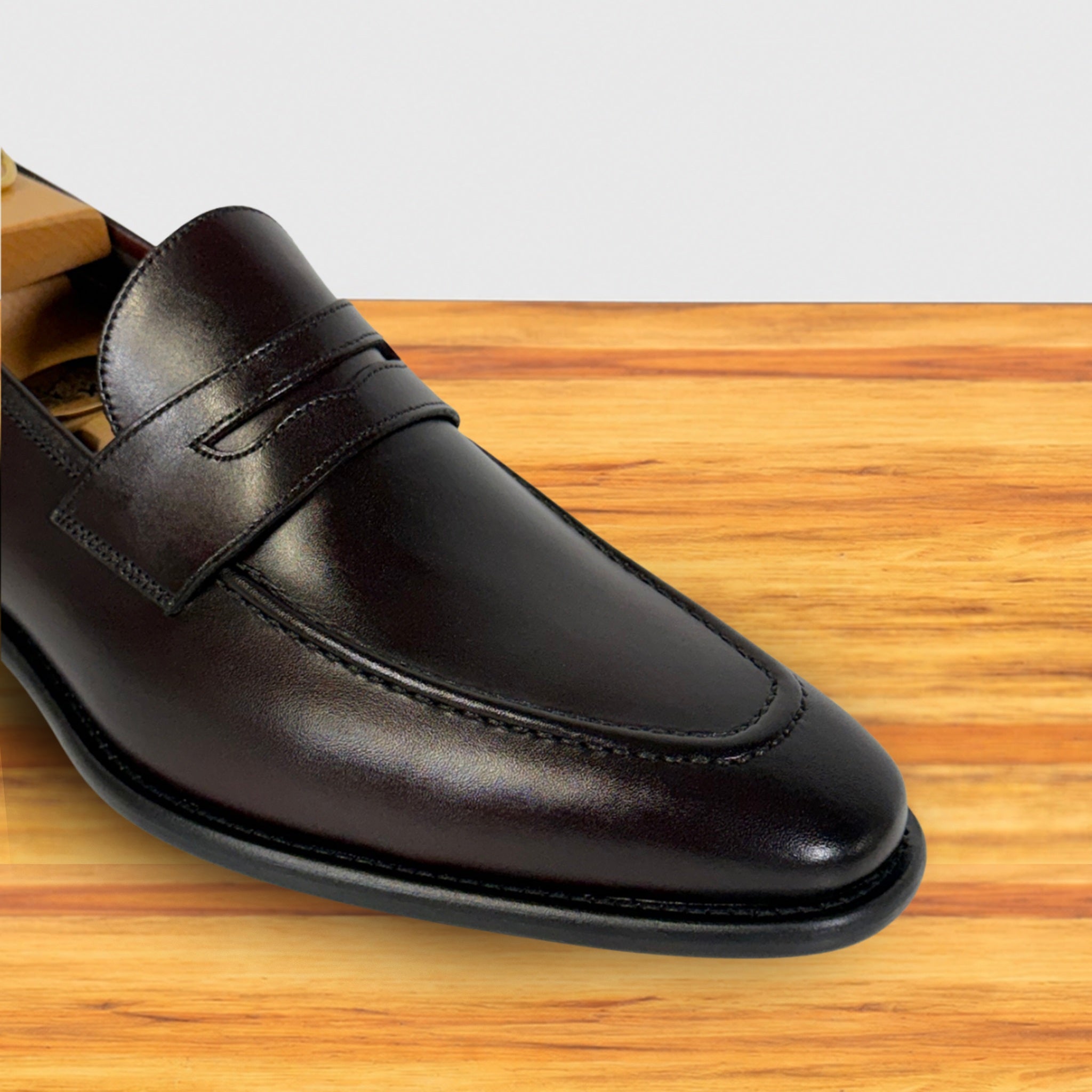 A Burgundy leather penny loafer shown from a close-up angle on a wooden surface with a light gray background.