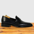 A Graphite leather penny loafer shoe with a pointed toe and low block heel, shown in profile on a wooden surface against a light gray background.