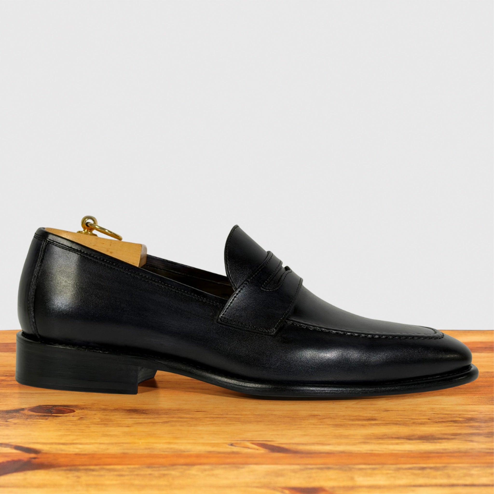 A Graphite leather penny loafer shoe with a pointed toe and low block heel, shown in profile on a wooden surface against a light gray background.