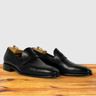 A pair of Graphite leather penny loafers with a low block heel, on a wooden surface against a light gray background.