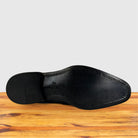 The outsole of a Graphite leather shoe is shown from a top-down view, resting on a wooden surface against a light gray background.