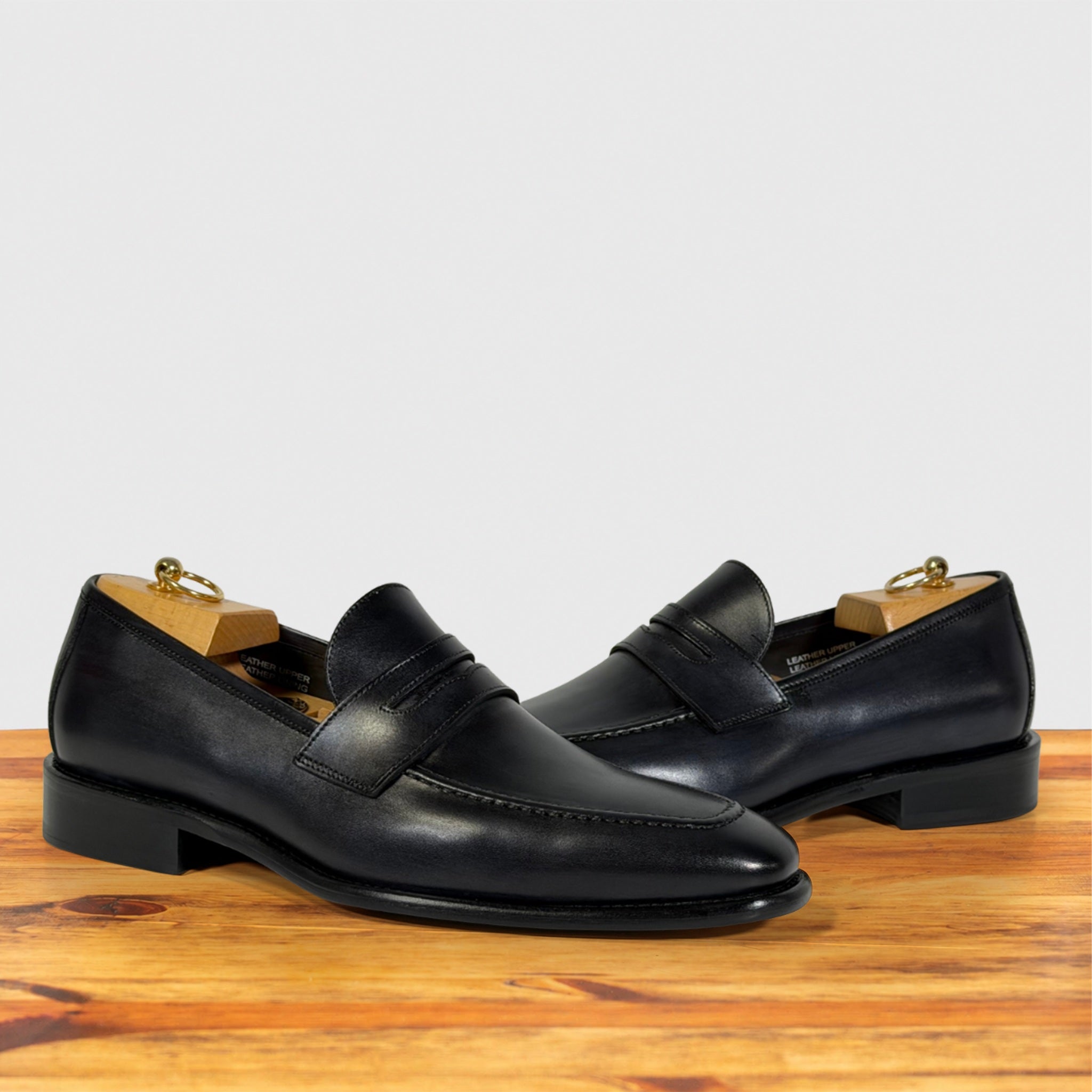 A pair of Graphite leather penny loafers , wooden shoe trees inside, displayed on a wooden surface against a light gray background.