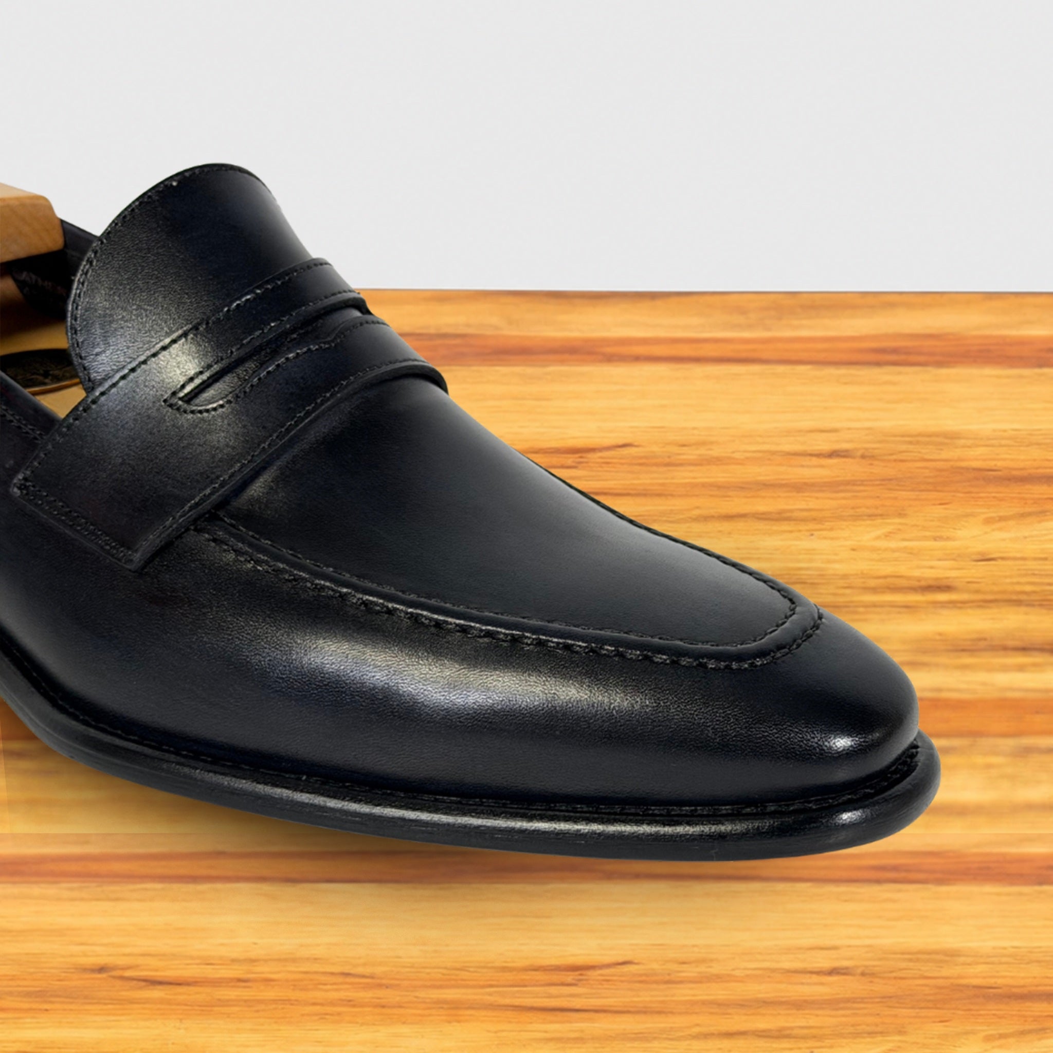 A Graphite leather penny loafer shoe shown from a side angle, resting on a light brown wooden surface against a white background.