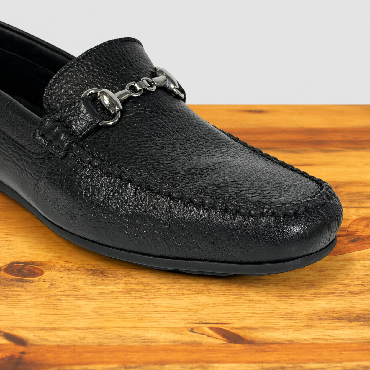 A black pebbled leather loafer with a silver buckle detail, shown on a wooden surface against a light gray background.