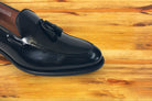A close-up of a black leather tassel loafer resting on a warm, light brown wooden surface.