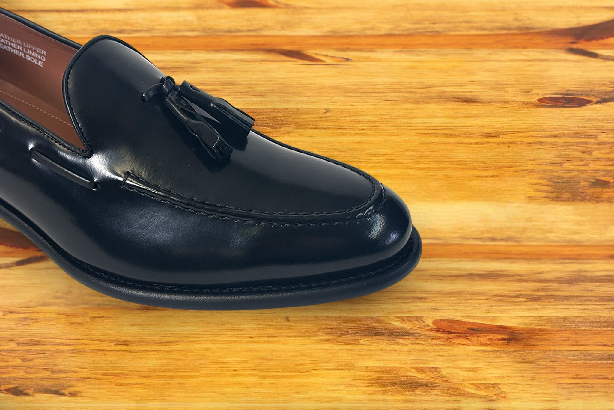 A close-up of a black leather tassel loafer resting on a warm, light brown wooden surface.