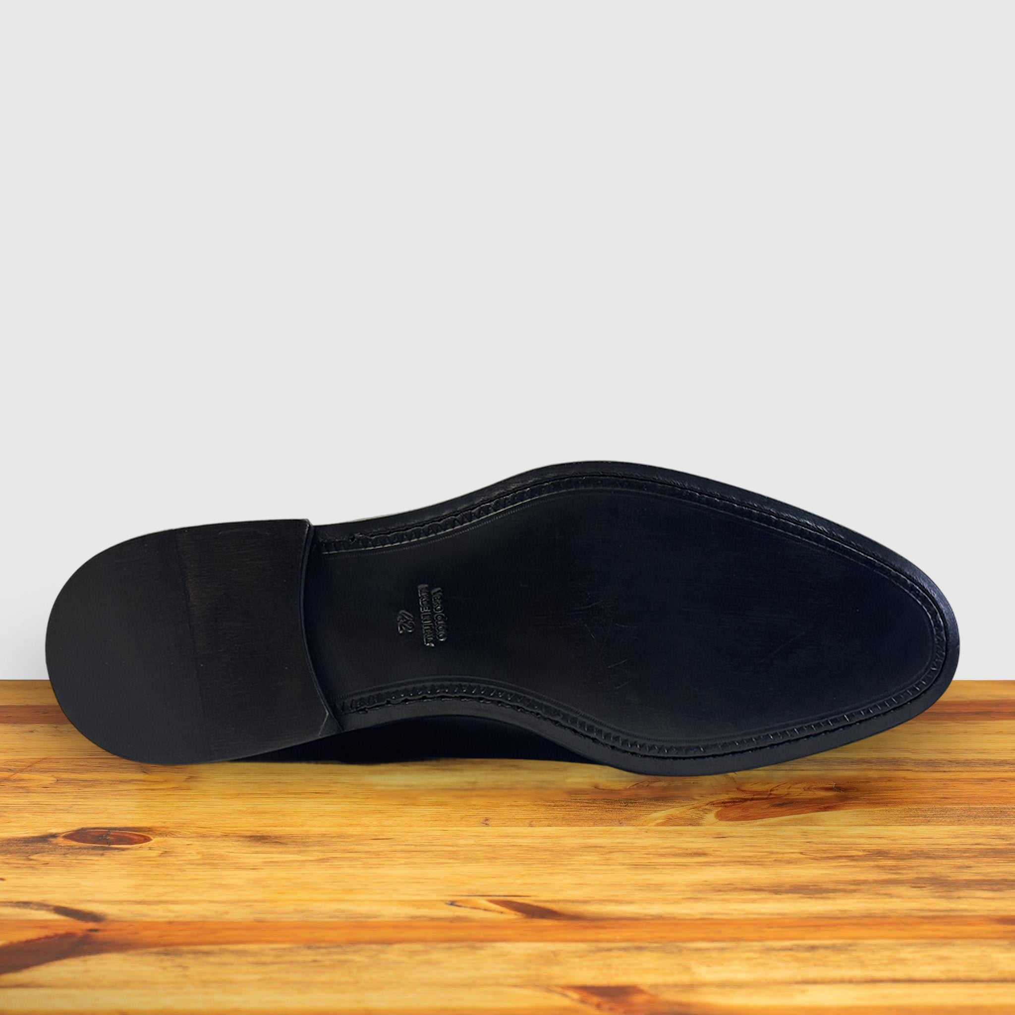 The outsole of a black leather dress shoe, showing the heel, toe, and center line stitching, resting on a wooden surface against a light gray background.