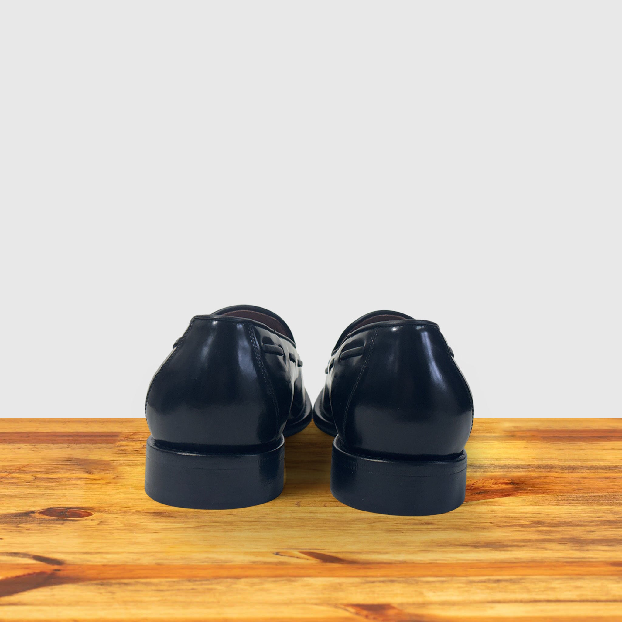 A pair of black leather dress shoes shown from the back, resting on a wooden surface against a light gray background.