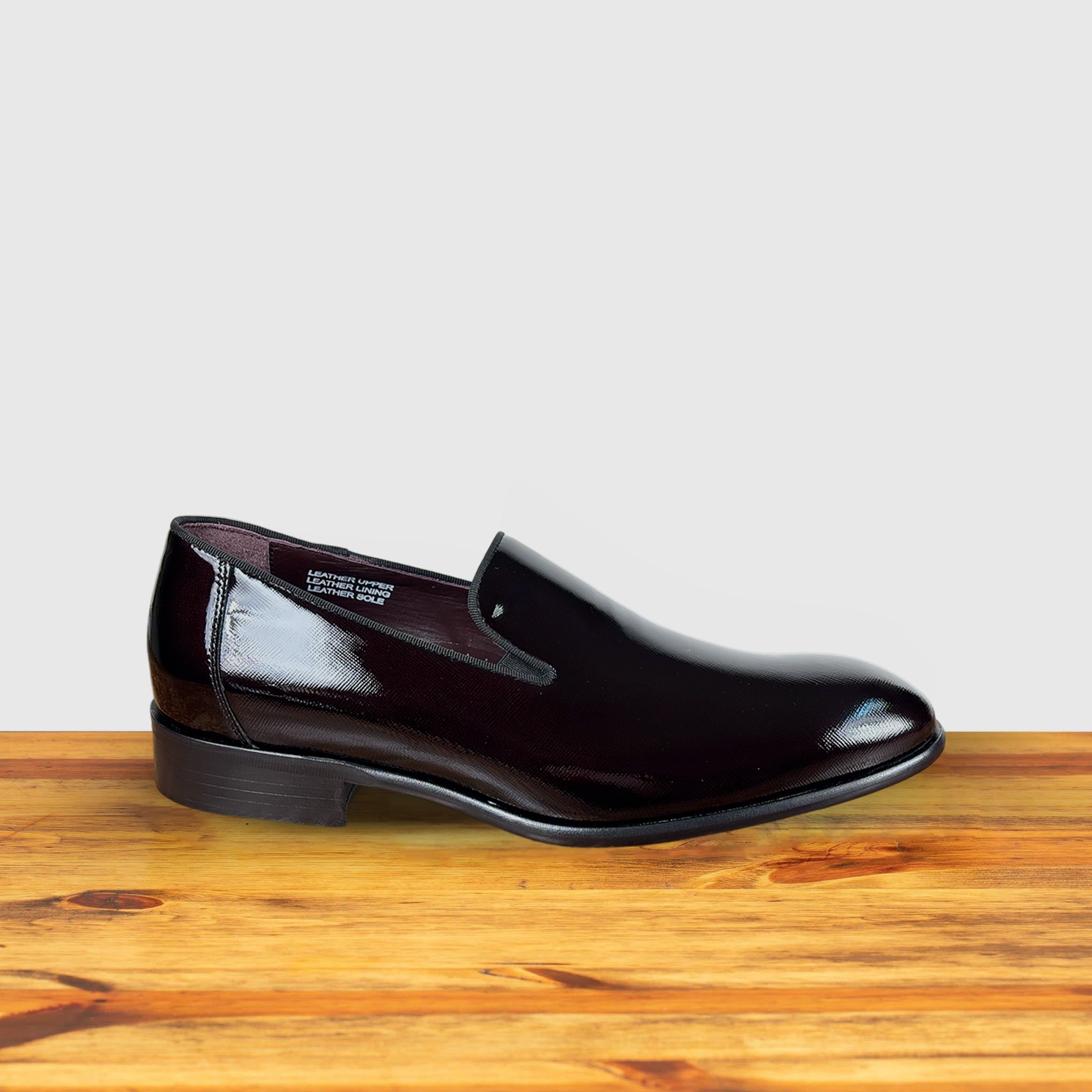 A single Burgundy patent leather slip-on shoe placed on a wooden surface, with a polished finish and a low heel.