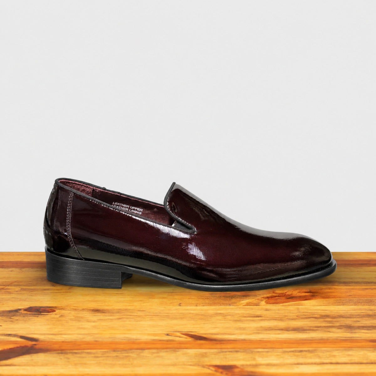 A Burgundy patent leather slip-on dress loafer with a black sole, shown in side profile on a wooden table