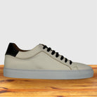 A low-top white and black sneaker with a light beige textured leather upper, black laces, a black heel tab, and a light gray sole, displayed on a wooden surface against a light gray background.