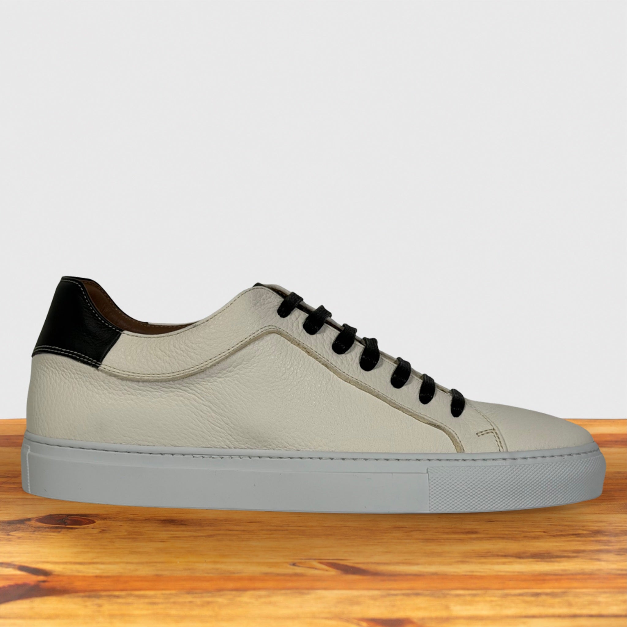 A low-top white and black sneaker with a light beige textured leather upper, black laces, a black heel tab, and a light gray sole, displayed on a wooden surface against a light gray background.