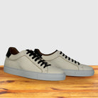A pair of low-top leather sneakers in white with black laces, black heel tab accents, and light gray soles, displayed on a wooden surface against a light gray background.
