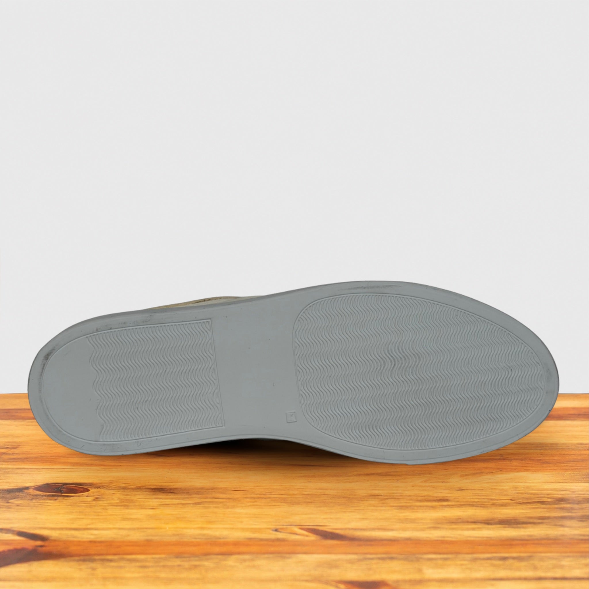 The bottom sole of a shoe, shown from a top-down view resting on a wooden surface against a light gray background.