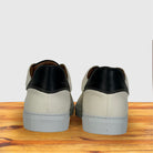 A pair of low-top sneakers shown from the back, featuring a black leather heel tab, white textured leather vamp, displayed on a wooden surface against a light gray background.