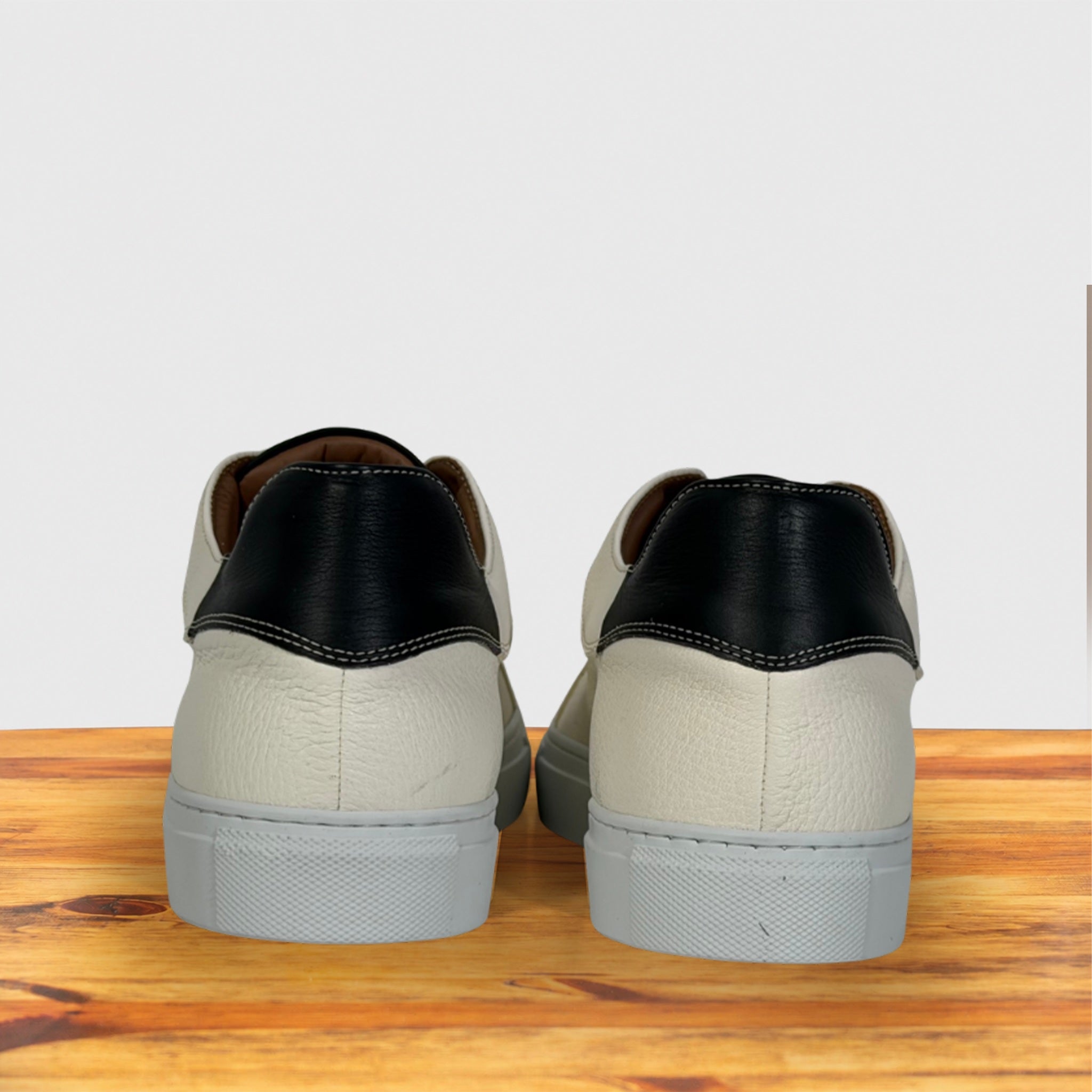 A pair of low-top sneakers shown from the back, featuring a black leather heel tab, white textured leather vamp, displayed on a wooden surface against a light gray background.