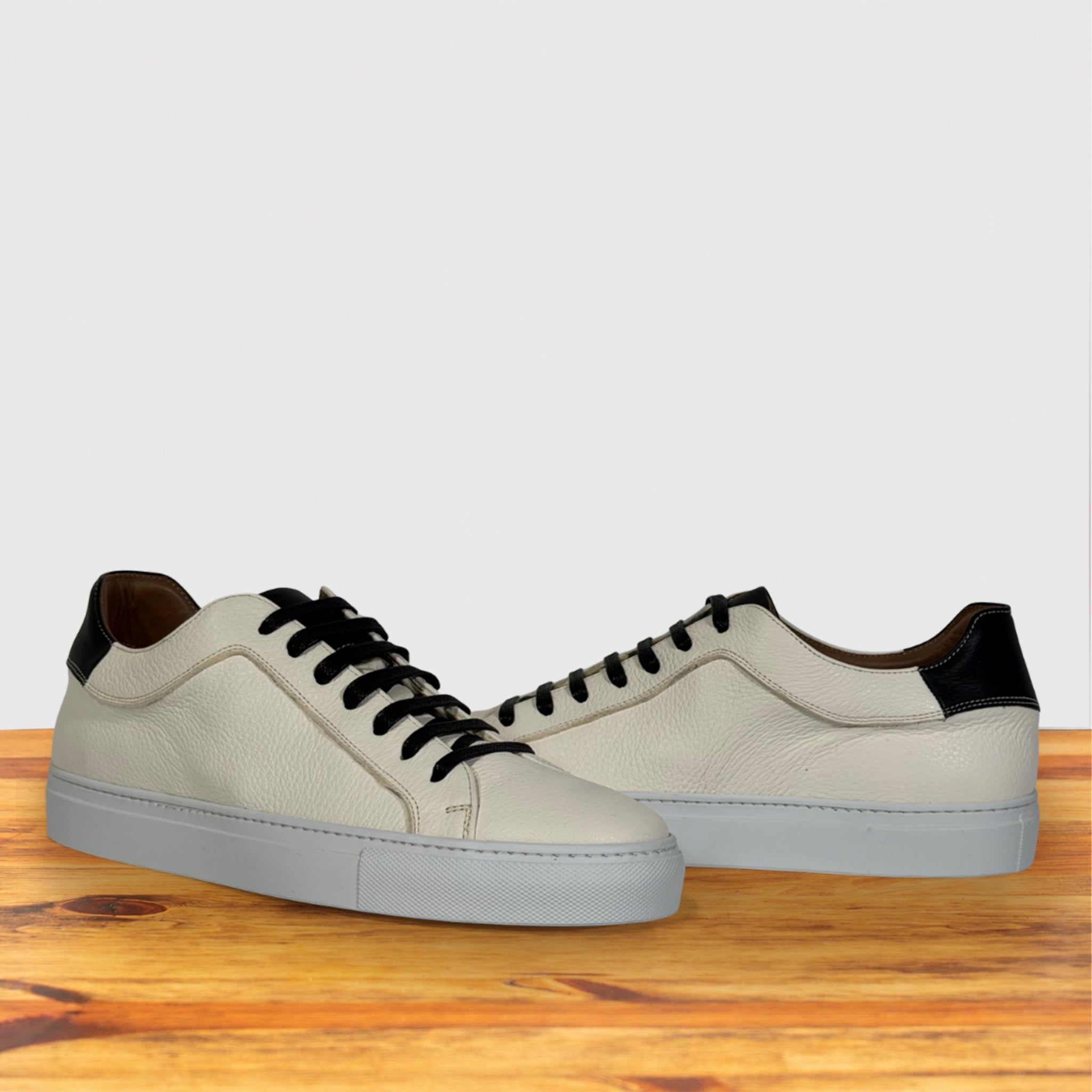 A pair of low-top leather sneakers in white with black laces, black heel tab accents, and light gray soles, displayed on a wooden surface against a light gray background.