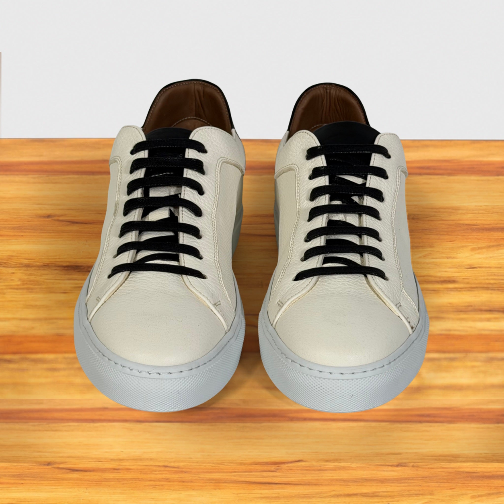 A pair of low-top sneakers with white leather uppers, black laces, and light gray soles, displayed on a wooden surface against a white background.