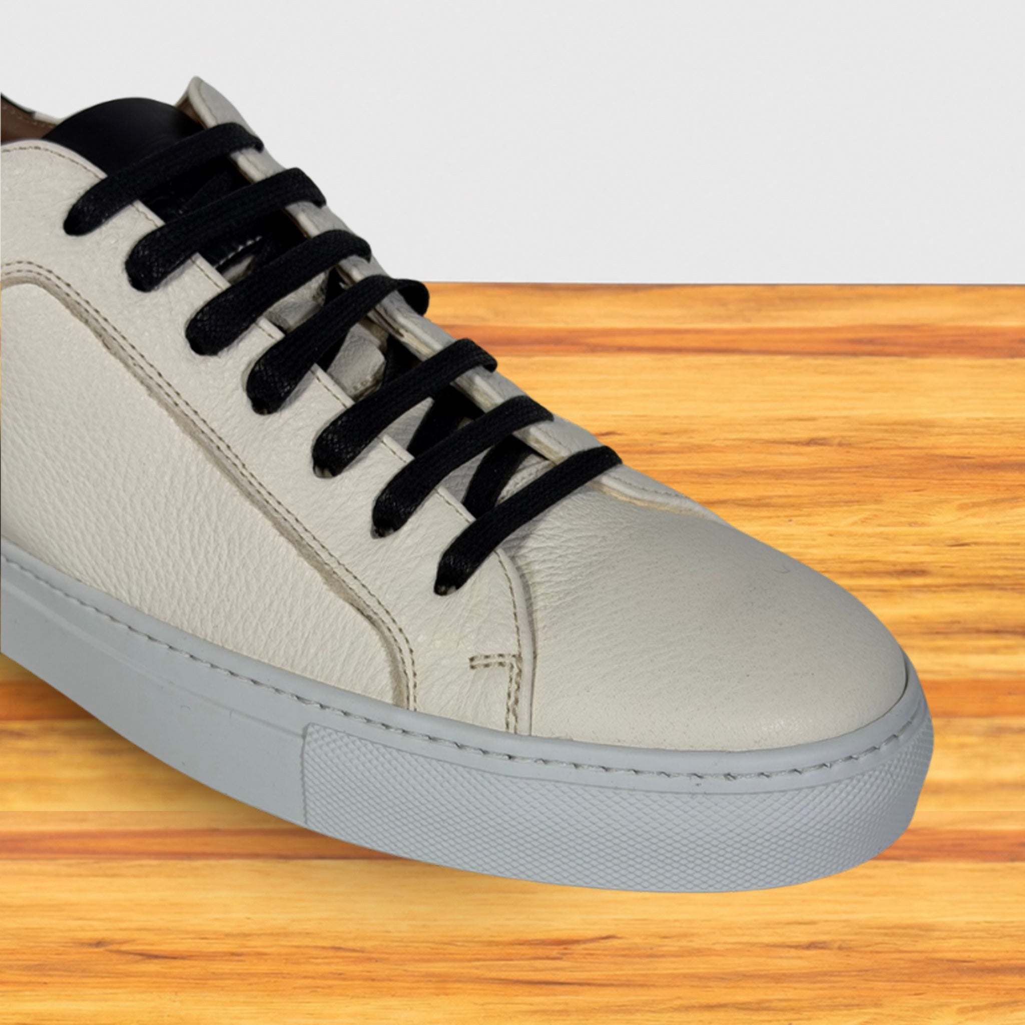 A white sneaker with black laces, and a light gray sole, shown on a wooden surface against a white background.
