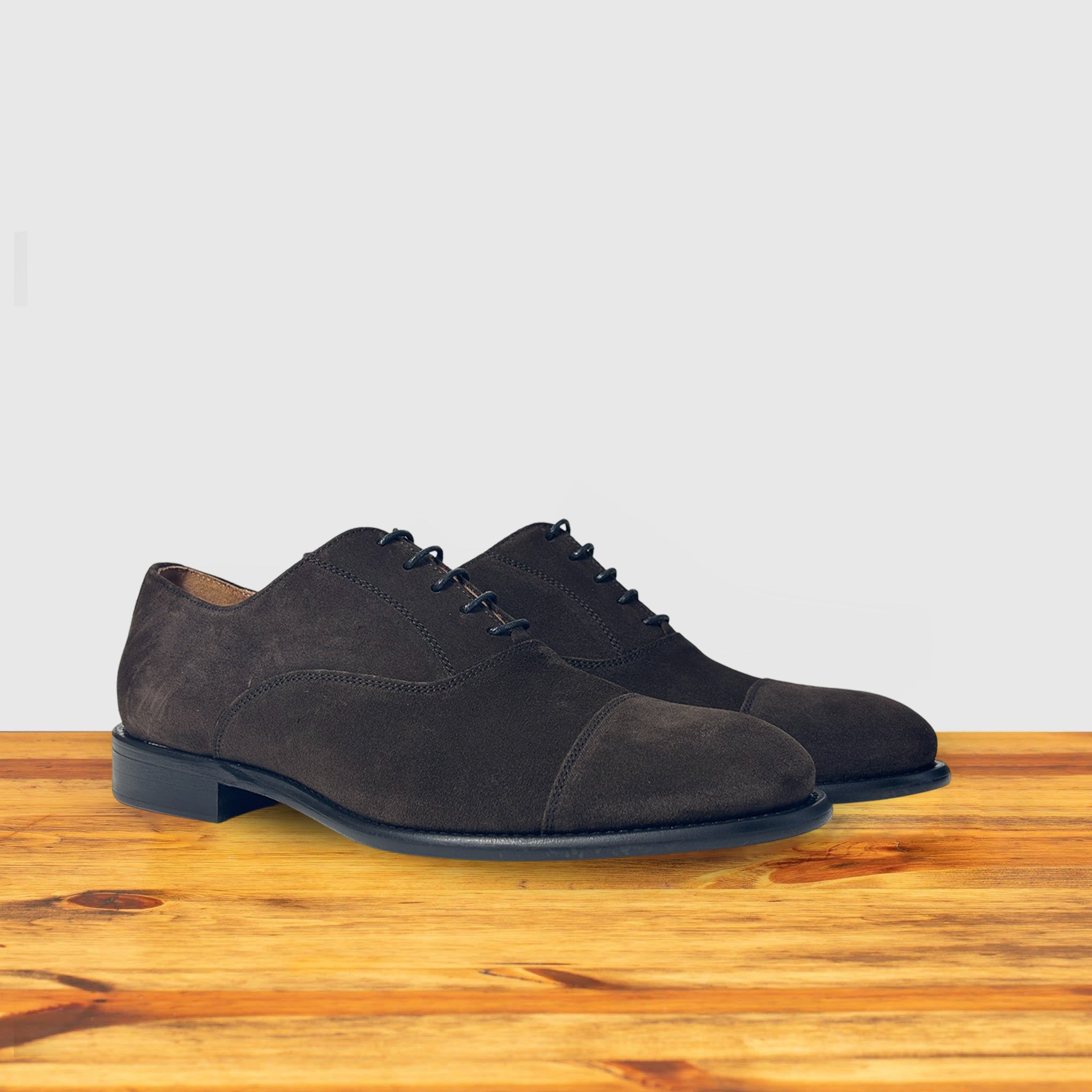 A Pair of Charcoal suede Oxford cap-toe shoe with a leather sole, displayed on a wooden surface.