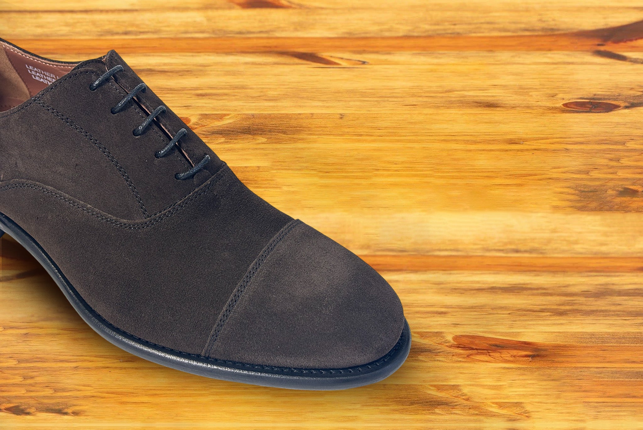 A Charcoal suede Oxford cap-toe shoe with a leather sole, displayed on a wooden surface.